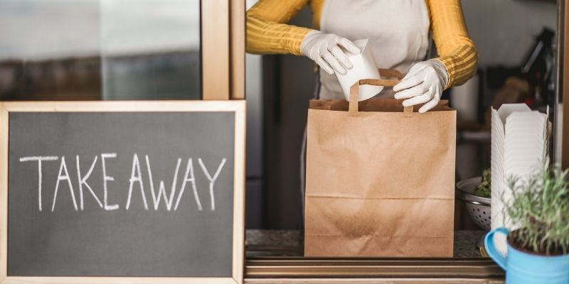 which-industries-most-impacted-covid-19 Female restauranteur packing takeaway meal into brown paper bag for pickup or delivery during COVID-19 pandemic